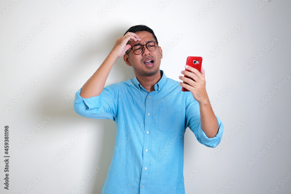 Adult Asian man showing disappointed expression while looking to his mobile phone Adult Asian man showing disappointed expression while looking to his mobile phone