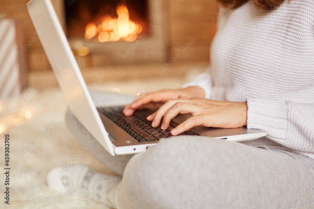 Fototapeta premium Faceless unknown woman sitting on floor on soft carpet at fireplace with laptop and working, typing, wearing white sweater and gray trousers, posing against burning fire on background.