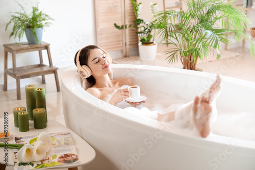 Young woman with headphones and cup of coffee taking bath at home