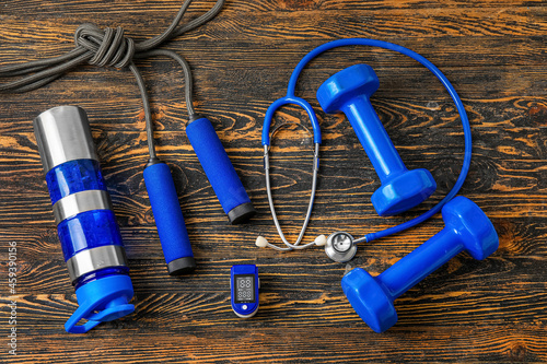 Composition with pulse oximeter, stethoscope, bottle of water and sport equipment on wooden background