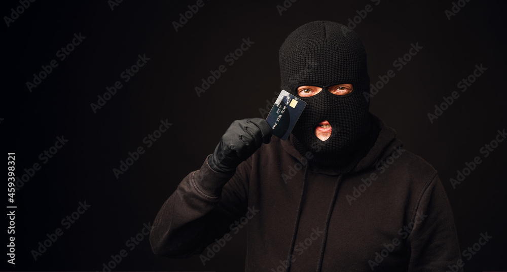 Thief in black balaclava holding black credit card. Banking and non cash thief concept