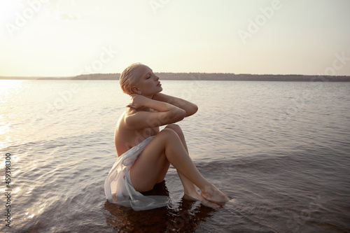 Art nude sexy blonde with short haircut is sitting in water on shore beach of lake at sunset. Wet hair and a woman body. Secluded beach holiday