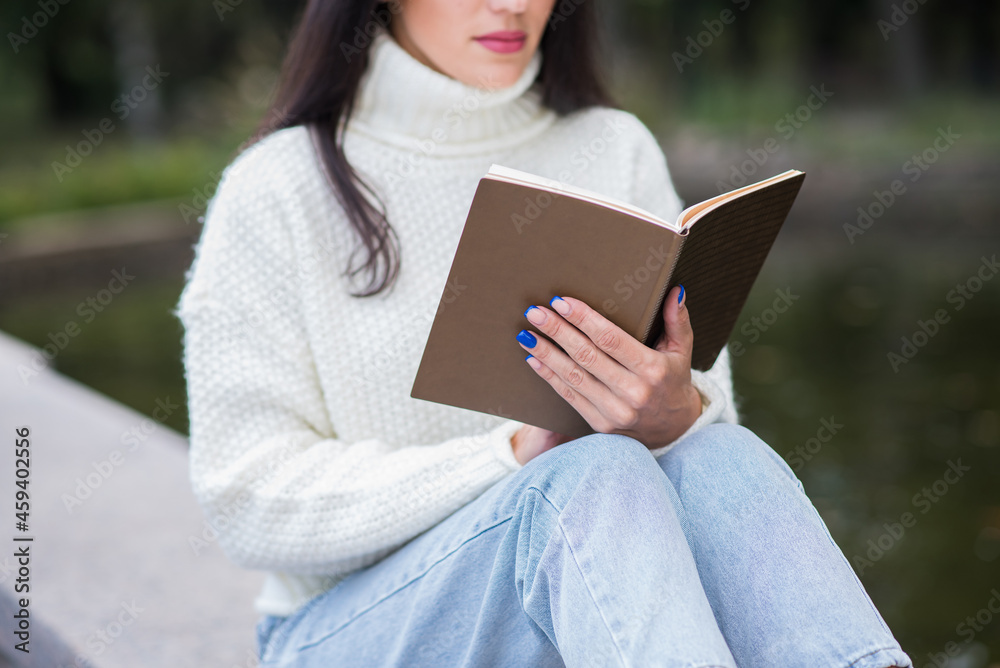 Obraz premium A young and attractive Caucasian brunette girl sitting and reading a book in a park near the river. The student is studying. The concept of education and knowledge.