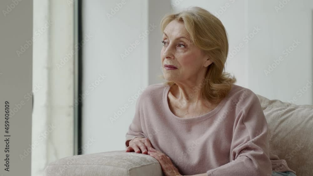 A thoughtful elderly woman is sitting on the sofa in the living room, looking out of the window into the distance, thinking or thinking A thoughtful elderly woman is sitting on the sofa in the living room, looking out of the window into the distance, thinking or thinking