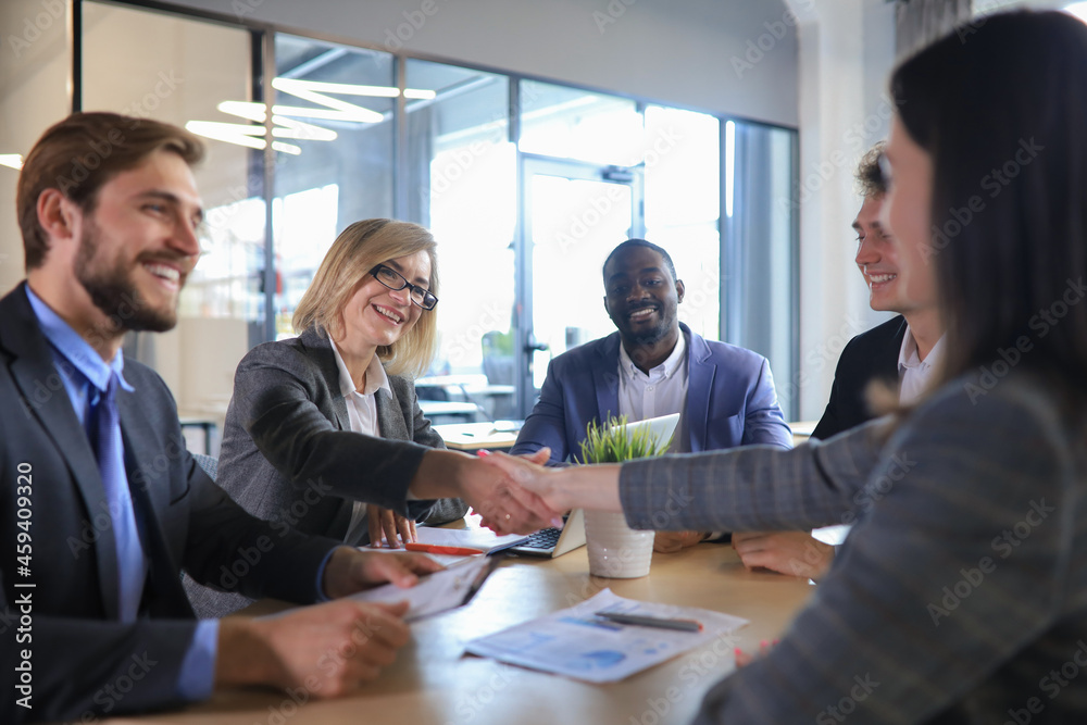 Two women shaking hands and looking at each other with smile while their coworkers sitting at the business meeting. Two women shaking hands and looking at each other with smile while their coworkers sitting at the business meeting.