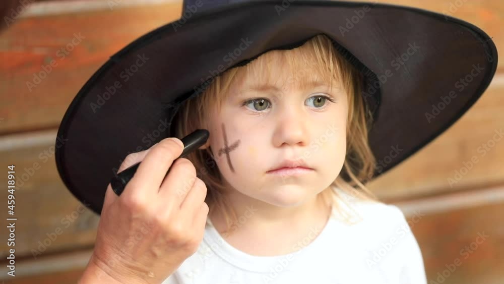 Little girl in black witch hat touches head by wooden house Little girl in black witch hat touches head by wooden house