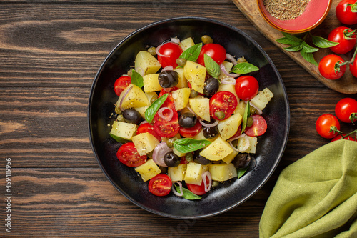 Vegetarian mediterranean salad. Made with boiled potatoes, tomatoes, onions, oregano, capers, black olives, basil leaves. Dark wooden background, top view.