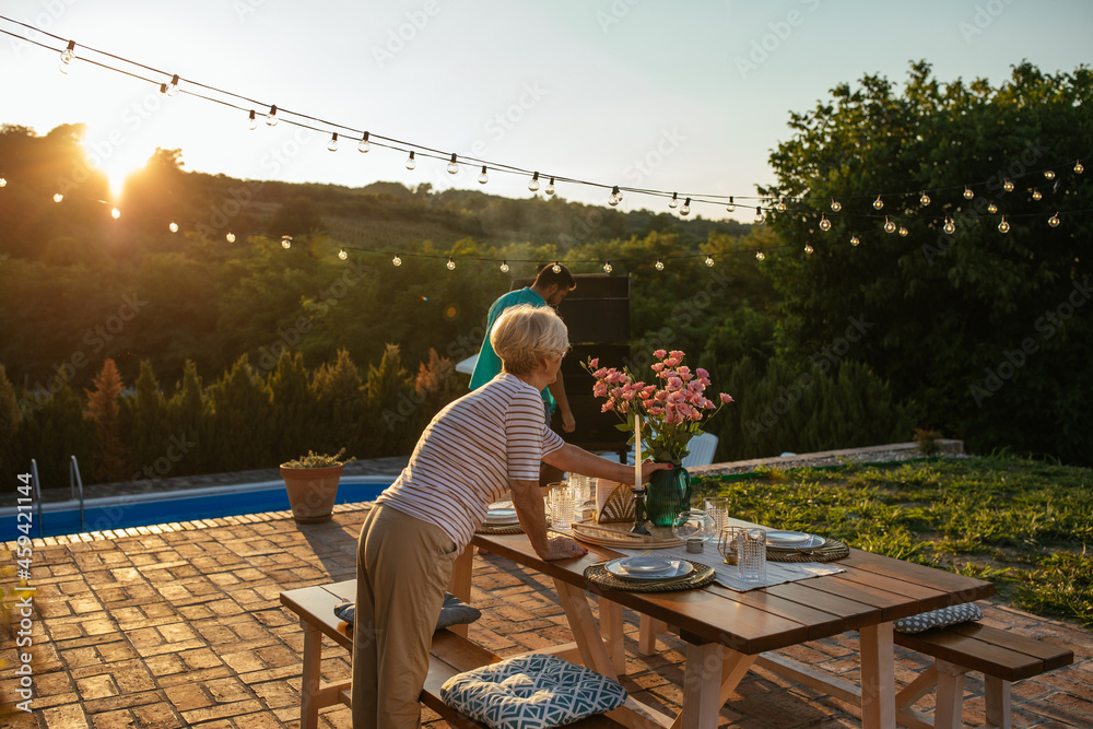 Senior woman setting dining table in the backyard and preparing everything for family gathering Senior woman setting dining table in the backyard and preparing everything for family gathering