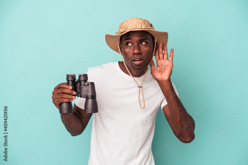Young African American man holding binoculars isolated on blue background trying to listening a gossip.