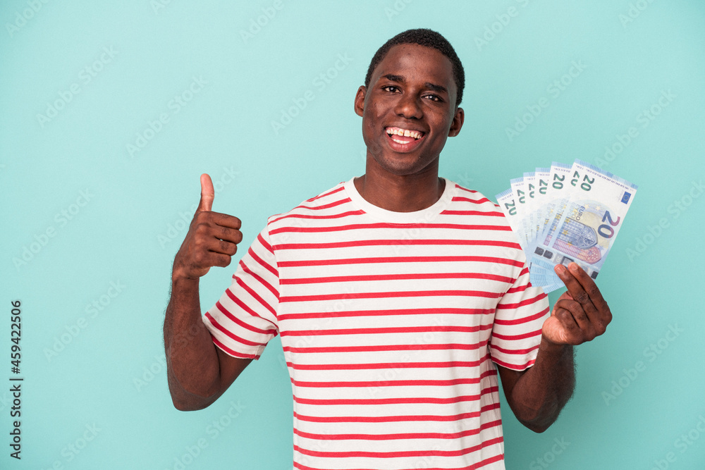 Young African American man holding a bank notes isolated on blue background smiling and raising thumb up Young African American man holding a bank notes isolated on blue background smiling and raising thumb up