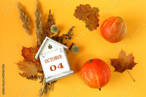 Calendar for October 4 : decorative house with the name of the month in English and the numbers 04 on bouquets of dried flowers, two orange pumpkins on a yellow background, top view photo