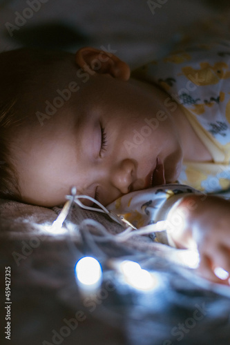 A charming baby sleeping in a cradle at night. A little boy in pajamas takes a nap in a dark room with a crib, in the lights of a garland. Sleeping time for children. 