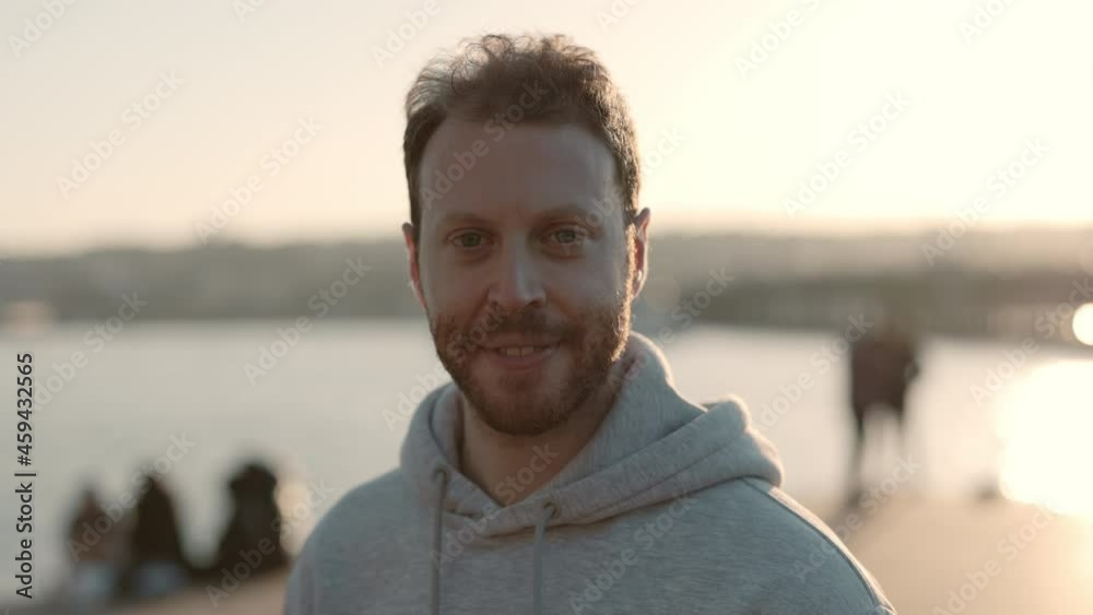 Portrait of redhead male runner in grey hoodie resting after evening run and looking at camera. Blur background of summer sunset over city river Portrait of redhead male runner in grey hoodie resting after evening run and looking at camera. Blur background of summer sunset over city river