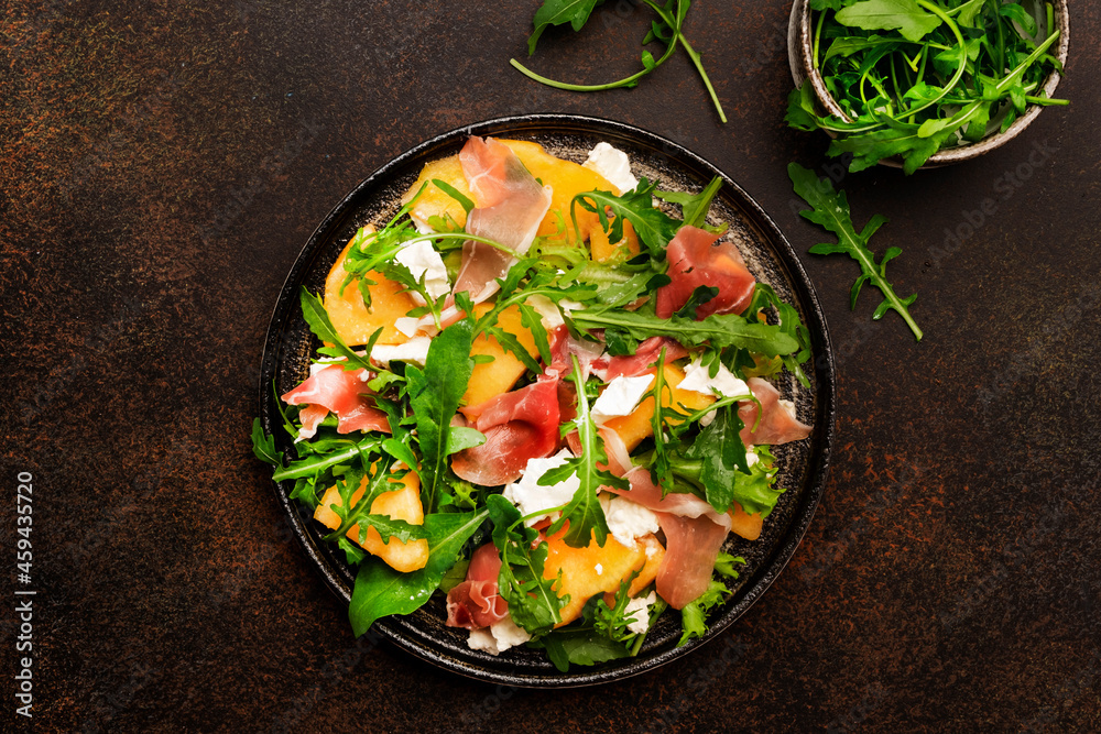 Salad with melon, jamon, goat cheese and arugula on brown background, top view, copy space