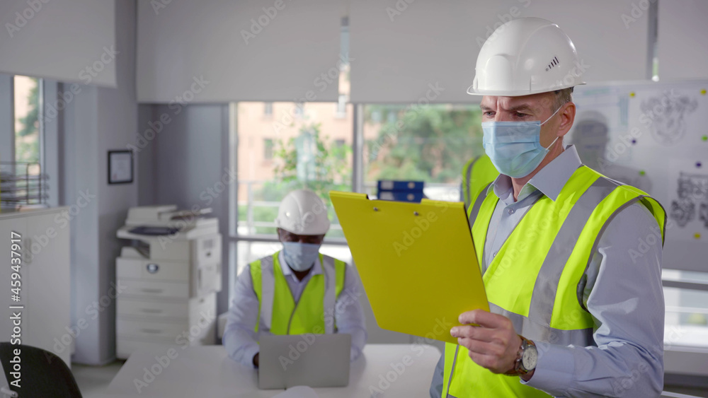 Mature engineer in safety mask reading document on clipboard in office Mature engineer in safety mask reading document on clipboard in office
