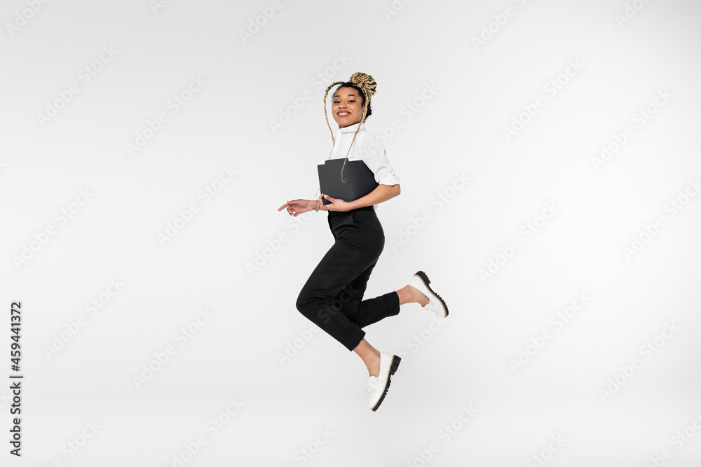 cheerful african american businesswoman looking at camera while levitating with paper folder isolated on white