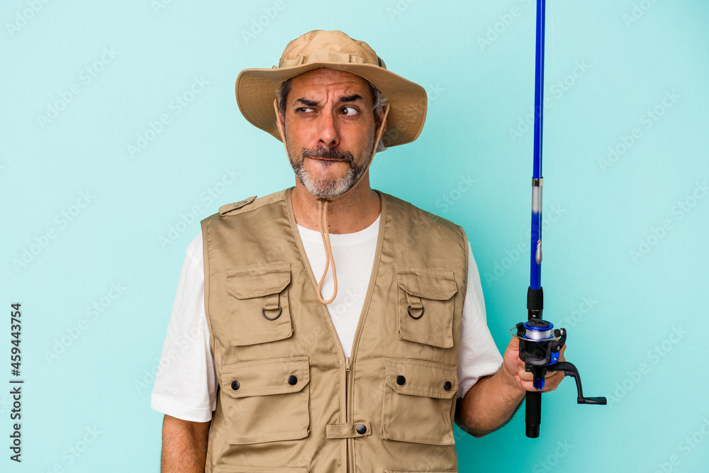 Middle age caucasian fisherman holding rod isolated on blue background confused, feels doubtful and unsure. Middle age caucasian fisherman holding rod isolated on blue background confused, feels doubtful and unsure.
