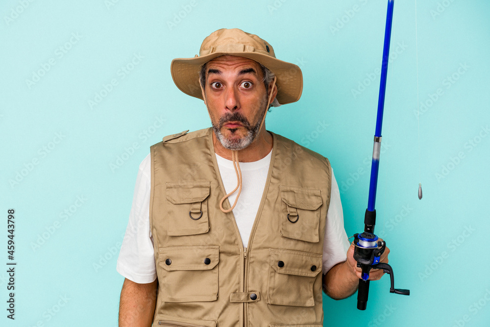 Middle age caucasian fisherman holding rod isolated on blue background shrugs shoulders and open eyes confused. Middle age caucasian fisherman holding rod isolated on blue background shrugs shoulders and open eyes confused.