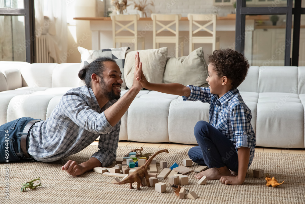 Happy African American father and son giving high five, celebrating success, playing with construction toys, wooden blocks, family spending leisure time together, sitting on warm floor in living room Happy African American father and son giving high five, celebrating success, playing with construction toys, wooden blocks, family spending leisure time together, sitting on warm floor in living room