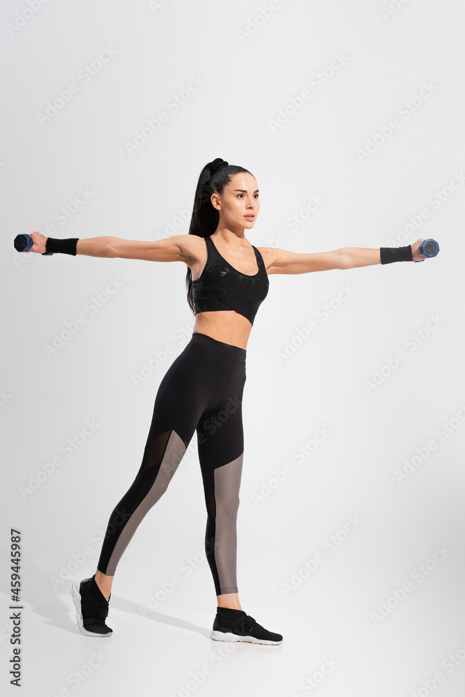 full length of strong young sportswoman in sportswear exercising with dumbbells on grey full length of strong young sportswoman in sportswear exercising with dumbbells on grey