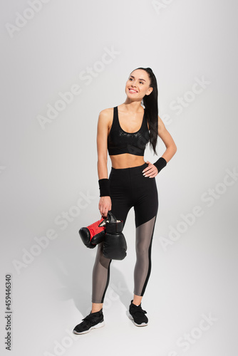 full length of happy sportswoman holding boxing gloves while posing with hand on hip on grey