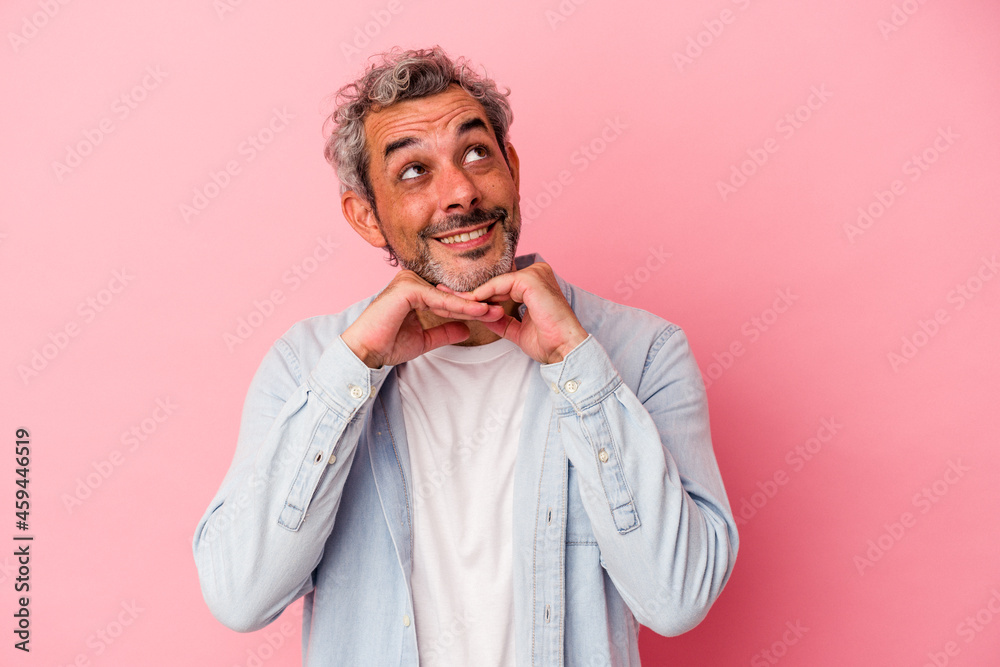 Middle age caucasian man isolated on pink background  keeps hands under chin, is looking happily aside.