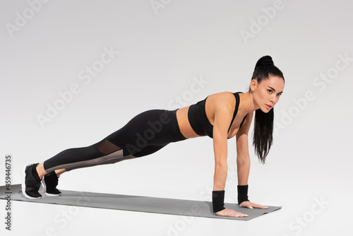 full length of young brunette woman in black sportswear doing plank on fitness mat on grey