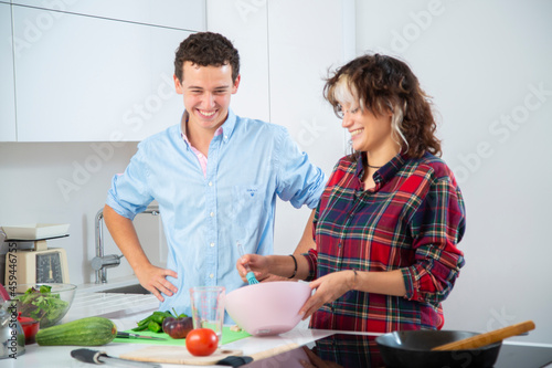 par jovenes sonrientes preparan una ensalada en un bol rosa vestidos con camisa azul y de cuadros en una cocina blanca de casa