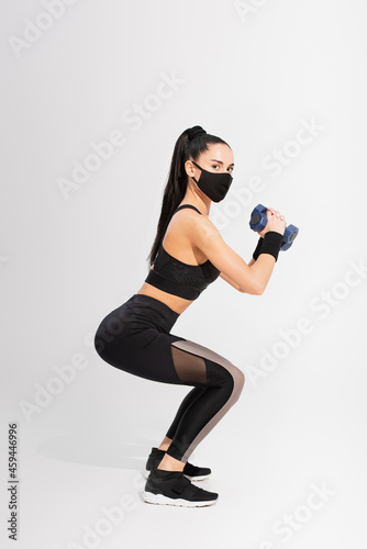 full length of sportswoman in black protective mask exercising with dumbbells on grey
