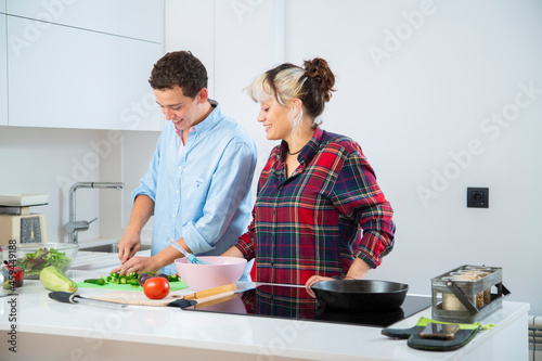 chico joven sonriente corta pimientos verdes con su pareja en una cocina blanca en la que hay mas verduras como tomate, calabacin y lechuga, utilizan una tabla de cortar, un bol rosa y una sarten