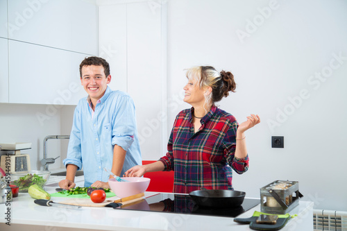 pareja joven sonriente cocina pimiento verde con una sarten de hierro en una cocina blanca con verduras, con placa de induccion, utilizan un bol rosa y usan herramientas de cocina