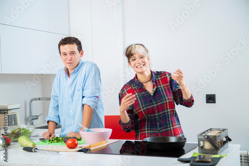 pareja joven sonriente cocina pimiento verde con una sarten de hierro en una cocina blanca con verduras, con placa de induccion, utilizan un bol rosa y usan herramientas de cocina