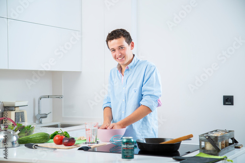 hombre joven sonriente hace una masa en un bol rosa, en una cocina blanca con placa de induccion, con una sarten de hierro y verduras, tomate, pimiento y usa herramientas de cocina