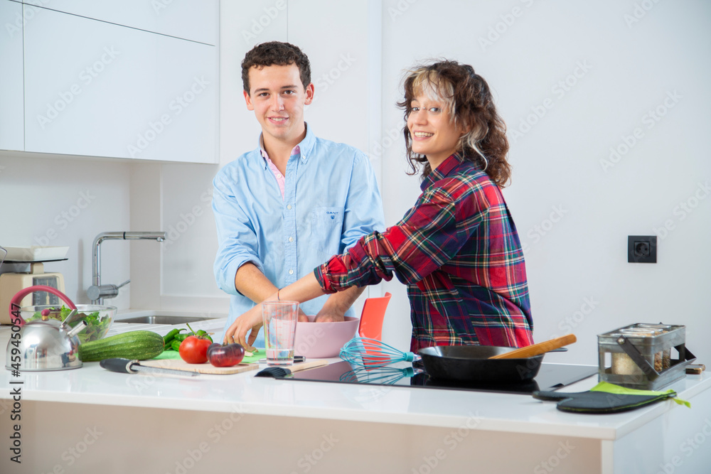 pareja joven sonriente cocinando verduras pimiento verde, tomate y calabacin, en una cocina blanca con placa de induccion, utilizan un bol rosa, una sarten de hierro y usan herramientas de madera