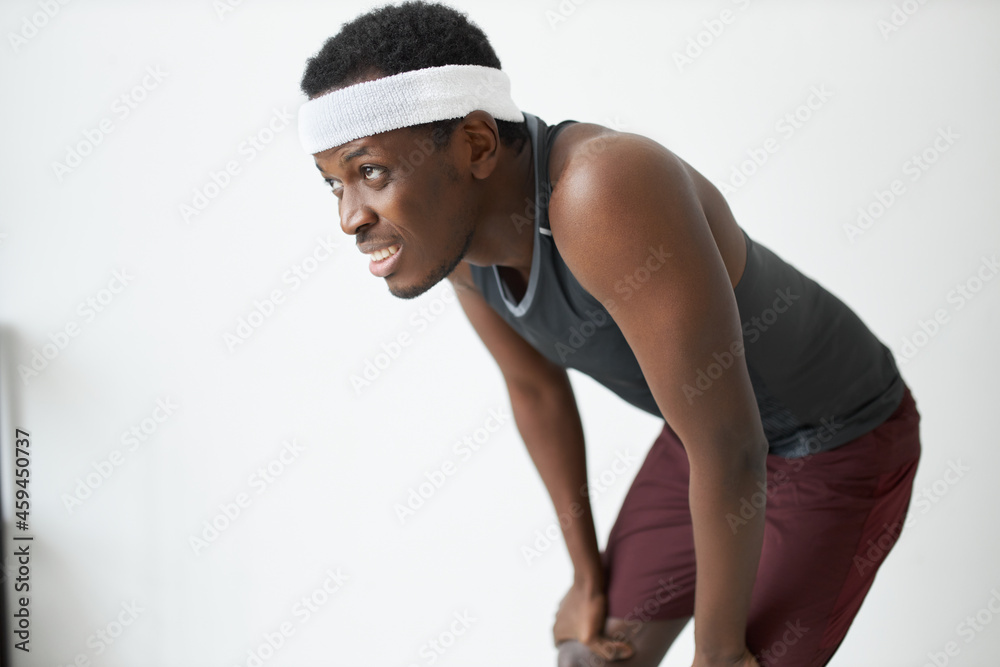 Ready, steady, go. Studio shot of young dark-skinned athlete in shorts, tank top and headband having rest after intensive workout, putting hands on half bent knees, looking in distance with tired face Ready, steady, go. Studio shot of young dark-skinned athlete in shorts, tank top and headband having rest after intensive workout, putting hands on half bent knees, looking in distance with tired face