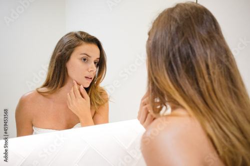 young white woman looking at her reflection in the mirror looking at skin looking for spots signs of age and lines serious and confident in the bathroom after shower self care skin care self love