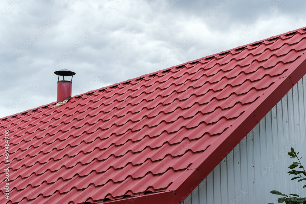 A roof made of red metal tiles with a chimney against a cloudy sky. A roof made of red metal tiles with a chimney against a cloudy sky.