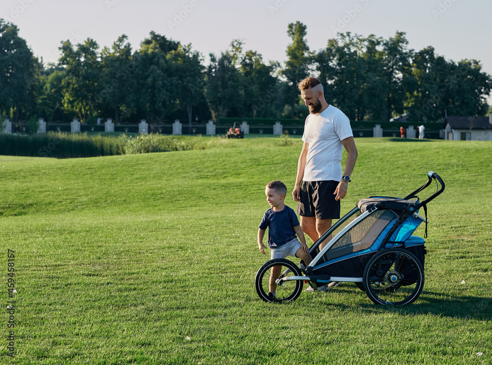 Father with his little son are walking in city park together on green grass with baby carriage. Fatherhood, daddy and child. Father's day Father with his little son are walking in city park together on green grass with baby carriage. Fatherhood, daddy and child. Father's day