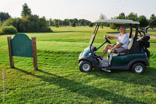 Golfer with prosthetic leg driving golf cart on grass course near golf club nameplate on a sunny day over landscape background and fairway