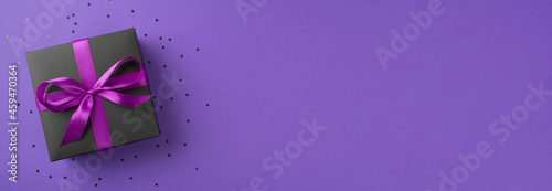 Top view photo of stylish black giftbox with violet ribbon bow and confetti on isolated violet background with copyspace