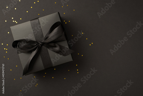 Top view photo of black giftbox with black ribbon bow and golden confetti on isolated black background with copyspace
