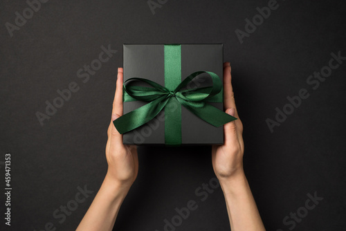 First person top view photo of female hands holding black giftbox with green satin ribbon bow on isolated black background