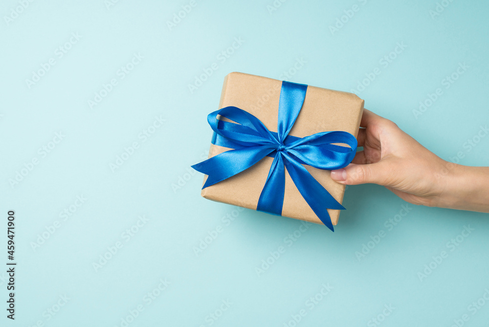 First person top view photo of female hand giving craft paper giftbox with vivid blue satin ribbon bow on isolated pastel blue background with empty space