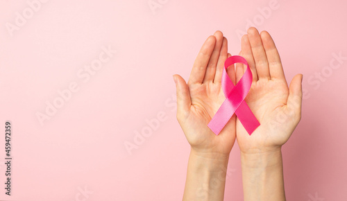 First person top panoramic view photo of woman's hands holding pink ribbon in palms symbol of breast cancer awareness on isolated pastel pink background with blank space