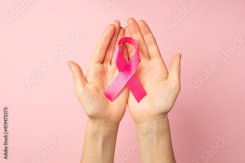 First person top view photo of hands holding pink ribbon in palms symbol of breast cancer awareness on isolated pastel pink background
