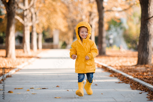 Pretty funny kid girl 2-3 year old wear yellow bright raincoat, rubber boots walk in park over fallen leaves outdoors. Autumn season. Happy child over nature background at city street.
