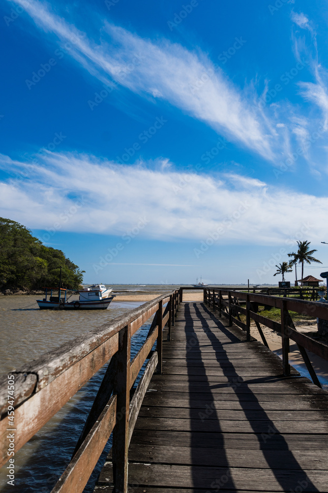 Fototapeta premium View of the beach of Rio das Ostras with the meeting of the river in Rio de Janeiro. Sunny day, blue sky. Yellow sand and some rocks. Wooden bridge to cross. Boats and fishing on the pier.