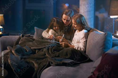 Beautiful caucasian woman reading fairy tale for two little girls while sitting together on cozy sofa. Mother and daughter covered with soft blanket using book before going to sleep.