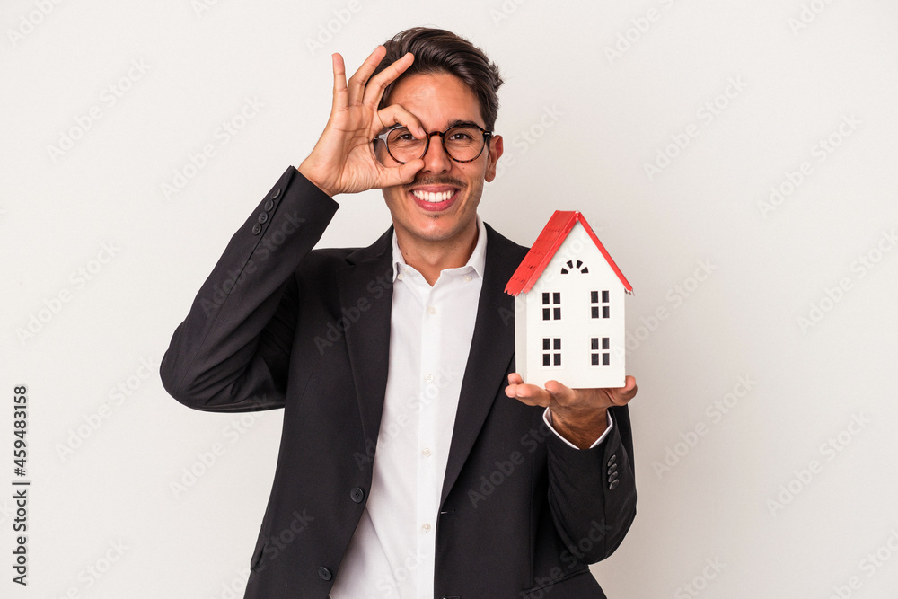 Young mixed race business man holding a toy house isolated on white background excited keeping ok gesture on eye. Young mixed race business man holding a toy house isolated on white background excited keeping ok gesture on eye.
