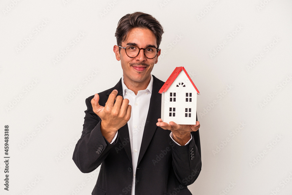 Young mixed race business man holding a toy house isolated on white background pointing with finger at you as if inviting come closer. Young mixed race business man holding a toy house isolated on white background pointing with finger at you as if inviting come closer.